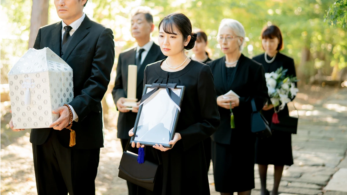 喪服姿のご遺族が遺影と白木の位牌、骨箱を胸に抱えて葬儀・告別式の日に会場へ進む様子