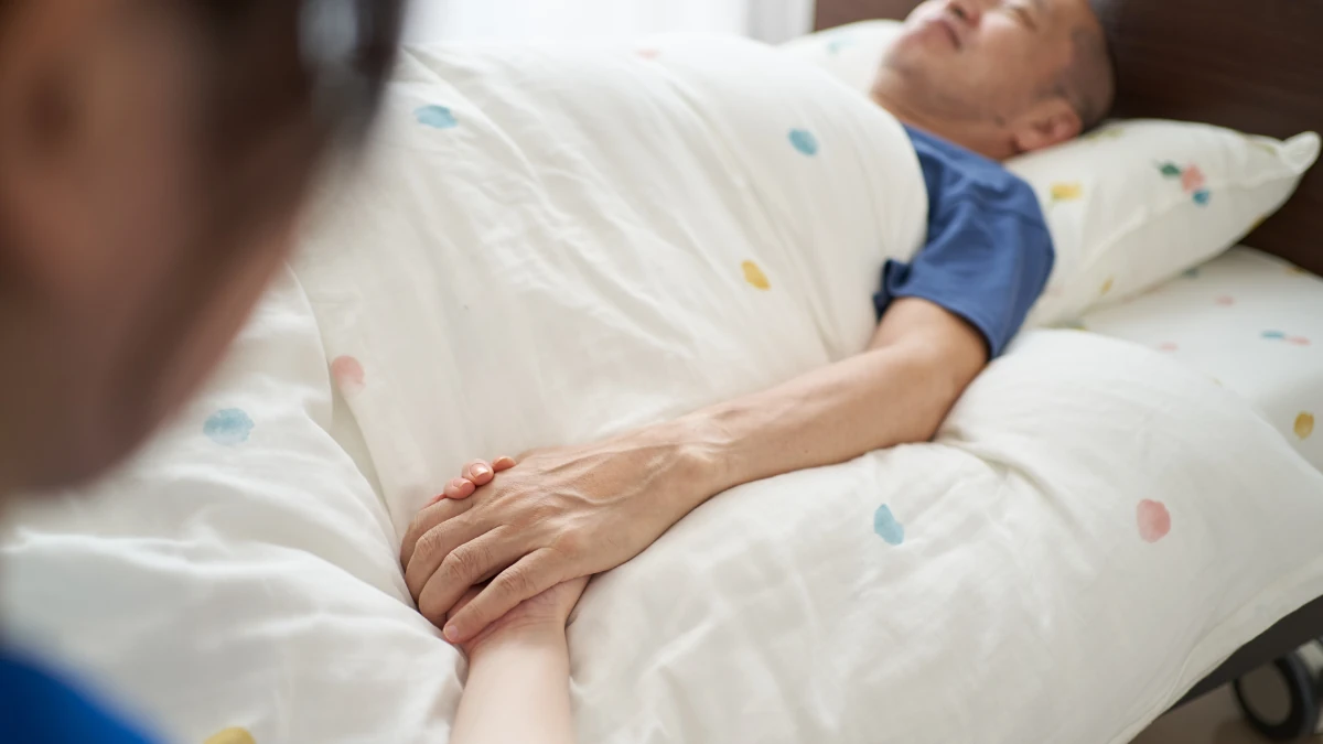 臨終を迎え病室のベッドに横たわる高齢男性の手を、家族がそっと握るご逝去直後の場面