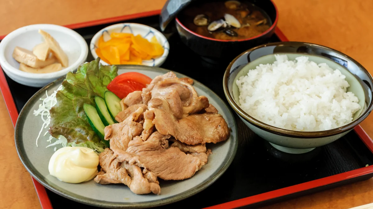 生姜焼きにご飯と味噌汁、小鉢と漬物が並んだ和定食を盆にのせた写真