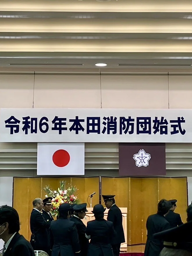 令和6年本田消防団始式の会場。金屏風の壇上に日の丸と区の旗が掲げられ、制服姿の消防団員が並んでいる