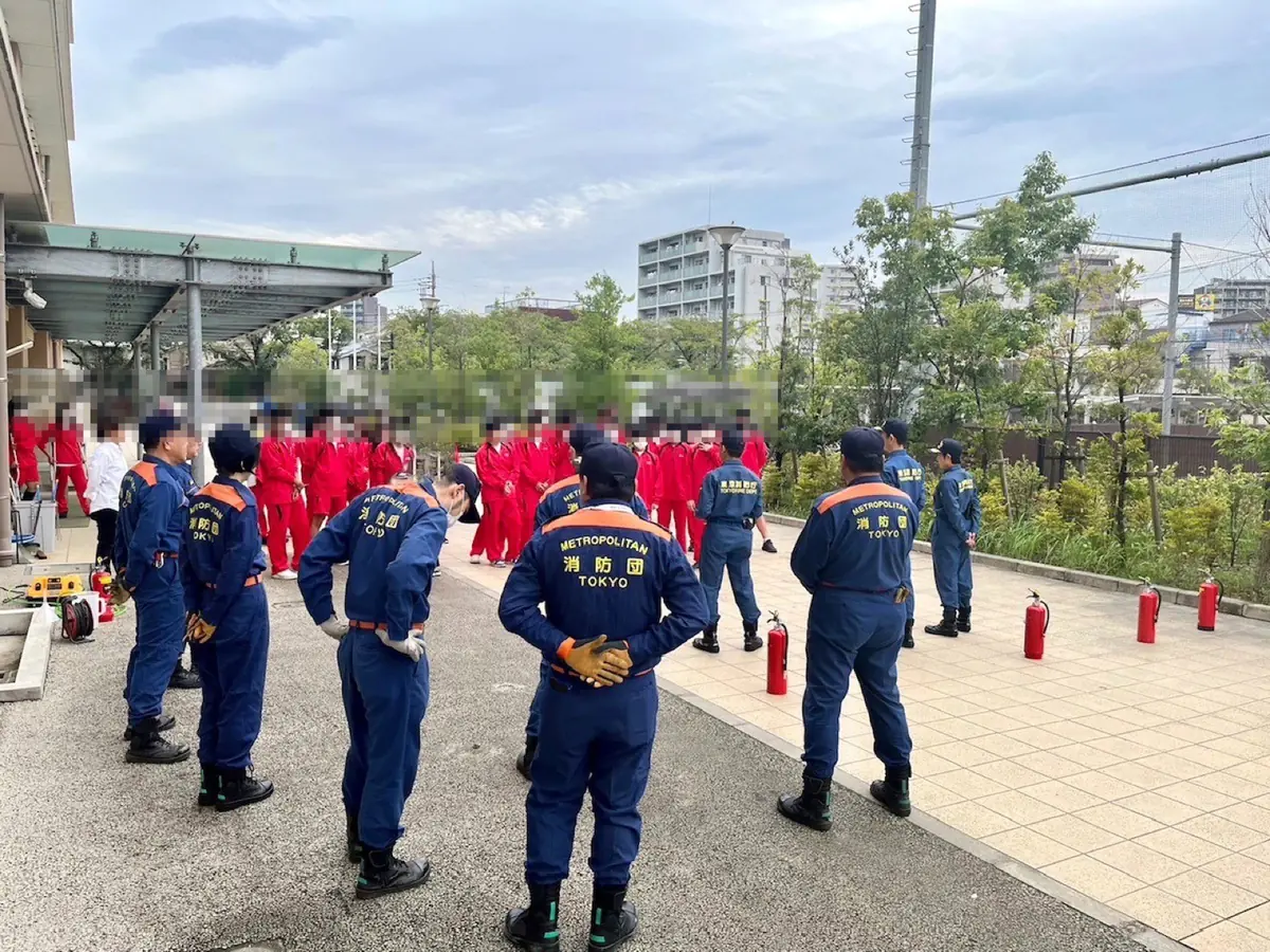 都立南葛飾高校の防災訓練。背中に「消防団」と書かれた制服の団員が並び、奥で赤いジャージの生徒たちが消火器の訓練を待っている