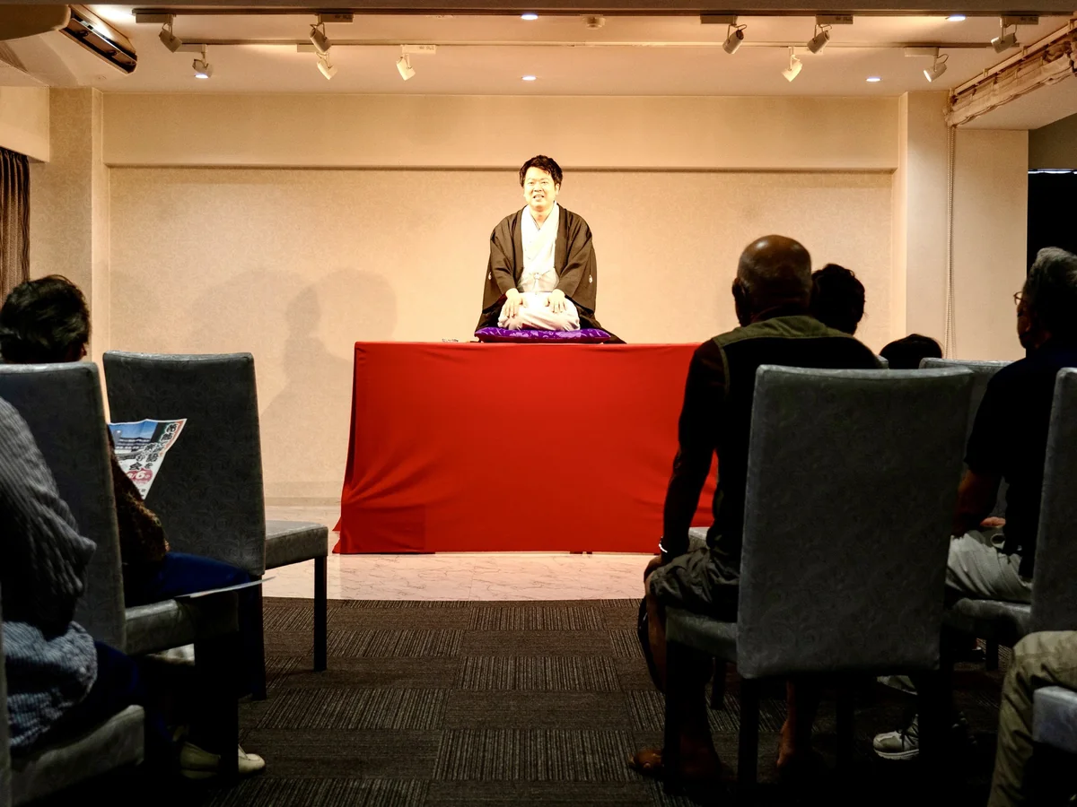第1回 葛飾で落語を楽しむ会の会場。赤い毛せんの高座に噺家さんが座り、手前の客席の皆さんが見守っている